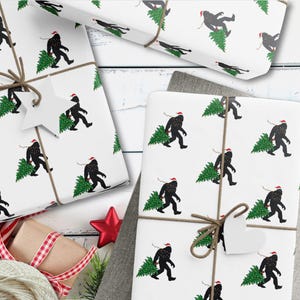 Puede incluir: Papel de regalo blanco con un patr&oacute;n repetitivo de siluetas negras de Bigfoot con gorros de Pap&aacute; Noel rojos y cargando &aacute;rboles de Navidad verdes. El papel est&aacute; atado con cordel marr&oacute;n y tiene una etiqueta de regalo en forma de estrella.
