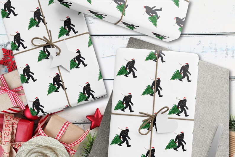Puede incluir: Papel de regalo blanco con un patr&oacute;n repetitivo de siluetas negras de Bigfoot con gorros de Pap&aacute; Noel rojos y cargando &aacute;rboles de Navidad verdes. El papel est&aacute; atado con cordel marr&oacute;n y tiene una etiqueta de regalo en forma de estrella.