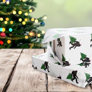 Puede incluir: Dos cajas de regalo rectangulares envueltas en papel blanco con un patr&oacute;n repetitivo de siluetas negras de Bigfoot cargando &aacute;rboles de Navidad verdes y usando peque&ntilde;os gorros de Pap&aacute; Noel rojos, apiladas sobre una superficie de madera.
