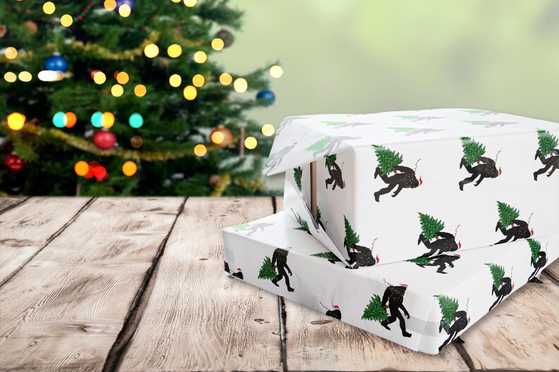 Puede incluir: Dos cajas de regalo rectangulares envueltas en papel blanco con un patr&oacute;n repetitivo de siluetas negras de Bigfoot cargando &aacute;rboles de Navidad verdes y usando peque&ntilde;os gorros de Pap&aacute; Noel rojos, apiladas sobre una superficie de madera.