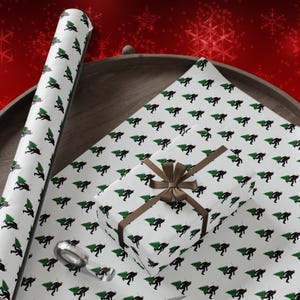 Puede incluir: Papel de regalo blanco con un patr&oacute;n repetitivo de siluetas negras de una criatura cargando un &aacute;rbol de Navidad verde. El conjunto incluye un tubo de papel enrollado, una hoja plana y una caja peque&ntilde;a envuelta con una cinta marr&oacute;n.