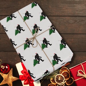 Puede incluir: Un regalo rectangular envuelto en papel blanco con un patr&oacute;n repetitivo de siluetas negras de Bigfoot con gorros de Pap&aacute; Noel rojos y cargando &aacute;rboles de Navidad verdes, asegurado con un cordel marr&oacute;n fino atado en un lazo.