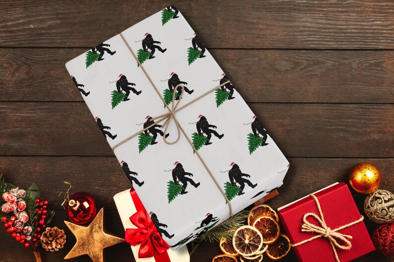 Puede incluir: Un regalo rectangular envuelto en papel blanco con un patr&oacute;n repetitivo de siluetas negras de Bigfoot con gorros de Pap&aacute; Noel rojos y cargando &aacute;rboles de Navidad verdes, asegurado con un cordel marr&oacute;n fino atado en un lazo.