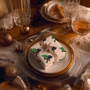 Puede incluir: Una caja de regalo cuadrada envuelta en papel blanco con un patr&oacute;n repetitivo de siluetas negras de una criatura cargando un &aacute;rbol de Navidad verde, rematada con una cinta de estrella dorada brillante, centrada en un plato blanco con borde dorado.