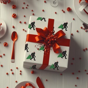Puede incluir: Una caja de regalo cuadrada envuelta en papel blanco con un patr&oacute;n repetitivo de siluetas negras de Bigfoot con un gorro de Pap&aacute; Noel rojo y cargando un &aacute;rbol de Navidad verde. La caja est&aacute; atada con una cinta roja brillante y un lazo grande.