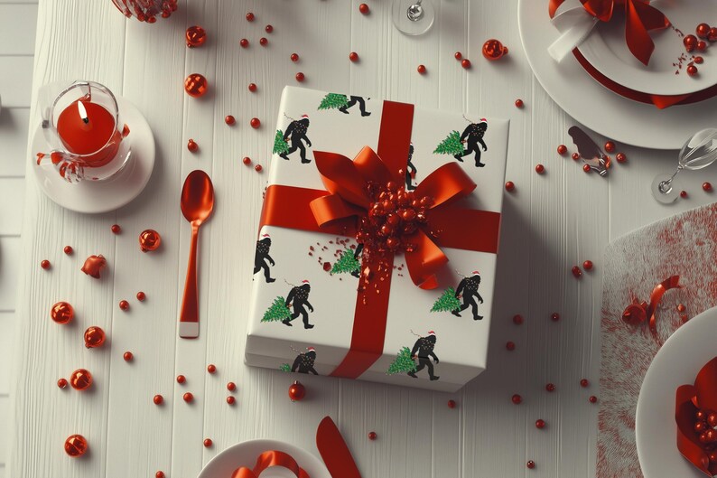 Puede incluir: Una caja de regalo cuadrada envuelta en papel blanco con un patr&oacute;n repetitivo de siluetas negras de Bigfoot con un gorro de Pap&aacute; Noel rojo y cargando un &aacute;rbol de Navidad verde. La caja est&aacute; atada con una cinta roja brillante y un lazo grande.