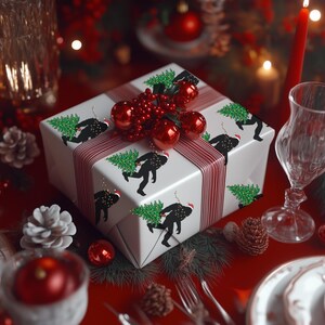 Puede incluir: Una caja de regalo cuadrada envuelta en papel blanco con un patr&oacute;n repetitivo de siluetas negras de una criatura cargando un &aacute;rbol de Navidad verde. La caja est&aacute; atada con una cinta a rayas rojas y blancas y decorada con adornos rojos.