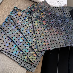 May include: A stack of six black pegboards with iridescent holographic patterns. The pegboards are made of a durable plastic material and have a grid of evenly spaced holes.