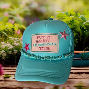 Peut inclure: Casquette de camionneur turquoise avec un dos en filet et une visière incurvée. L'avant présente un écusson avec le texte "PUT IT ON MY HUSBAND'S TAB" et deux étoiles roses. Une bordure décorative de petites pierres turquoises orne la visière.
