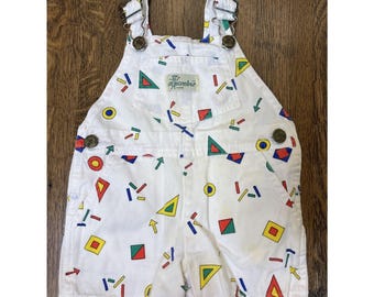 May include: White denim short dungarees featuring a colourful geometric print with red, blue, yellow, and green shapes including triangles, squares, circles, and rectangles. The garment has metal buckles on the straps and a front chest pocket with a label.