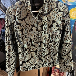 May include: A black jacket with a paisley pattern in gold and teal. The jacket has a mandarin collar and is buttoned up the front. The label reads "Jessica Collection". The jacket is hanging on a white hanger.