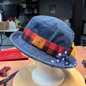 May include: Denim bucket hat with a red and black plaid band and a blue star-patterned accent. The hat features a brown leather patch with the word "ARCADE" and the text "EST. 2010".