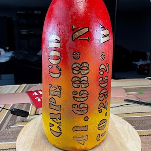 May include: A red, yellow, and orange ceramic buoy with the text "CAPE COD" and coordinates "41.6888° N 70.2962° W" painted on it.