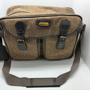 May include: A brown and black tweed camera bag with two front pockets, black leather accents, and a shoulder strap. The bag has a rectangular shape with a top flap and a tag on the side. The text "Leisure" is visible on the front.