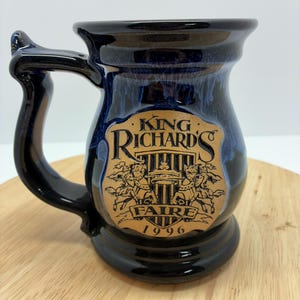 May include: A ceramic mug with a black handle and base, and a dark blue exterior with a glossy finish. The mug features a brown emblem with the words "King Richard's Faire 1996".