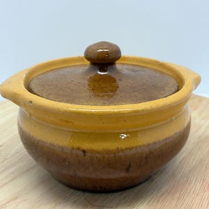 May include: A small, round ceramic pot with a lid. The pot is a warm brown color with a yellow rim and a matching lid. The lid has a small, rounded handle. The pot is sitting on a wooden surface.