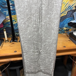 May include: A light gray scarf with a paisley pattern. The scarf is rectangular and has a border along the bottom edge. The scarf is hanging on a black metal hanger. The scarf has a fringe along the bottom edge.