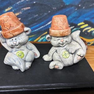 May include: Two ceramic figurines of gray cats wearing brown flower pot hats. Each cat has a green and yellow emblem on its belly. One cat is resting its head on its hand, while the other is sitting with its eyes closed.