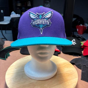 May include: A purple and teal Charlotte Hornets baseball cap. The cap features the team's logo on the front, with a teal brim and a teal top button. The cap is displayed on a white mannequin head.