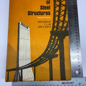 May include: A hardcover book titled Design of Steel Structures Second Edition by Boris Bresler, T. Y. Lin, and John B. Scalzi. The orange cover features a black graphic of a bridge structure. A metal ruler shows the book is 9.5 inches tall.