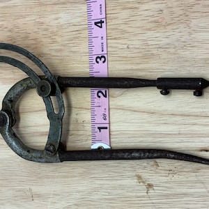 May include: An antique metal marking gauge with a curved handle and a pointed end. The gauge is approximately 4 inches long and is placed next to a pink measuring tape on a wooden surface. The tool appears to be vintage.