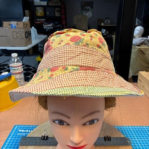 May include: A patchwork sun hat with a wide brim. The hat features a floral pattern, a red and white checkered pattern, and a green trim. The hat is displayed on a mannequin head.