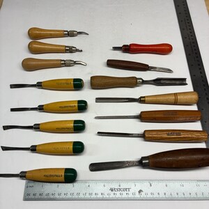 May include: A collection of fourteen woodworking tools including gouges and chisels with wooden handles, arranged alongside a Westcott ruler showing measurements in inches and centimeters to provide scale for the various metal blades and handles.