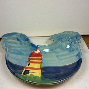 May include: A ceramic rooster-shaped serving dish with a blue and green glaze. The dish features a painted scene of a yellow lighthouse with a red roof against a blue sky.