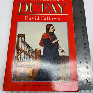 May include: A red paperback book titled The Master Musicians DUFAY by David Fallows, featuring an illustration of a person with a portative organ. A metal ruler shows the book is approximately 5.25 inches wide and 8.5 inches tall.