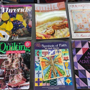 May include: A collection of quilting magazines, including "Threads," "Quilting," and "Symbols of Faith." The magazines feature colorful covers with images of quilts, patterns, and related crafts. The magazines are displayed on a gray surface.