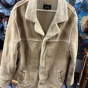 May include: A tan suede jacket with a cream-colored, fuzzy collar and lining. The jacket has a button-up front, two front pockets, and a label that reads "Patino". The jacket is hanging on a white hanger.
