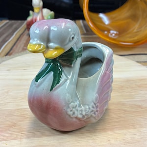 May include: A ceramic swan planter with a pink and white glaze. The swan has a green bow around its neck and a small flower design on its body. The planter has a small opening on the back for planting.