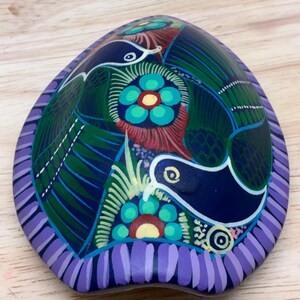 May include: Hand-painted, oval-shaped decorative stone with a vibrant design. The stone features a dark blue base with green, red, and yellow floral and bird motifs. The edges are bordered with a lavender and purple striped pattern.