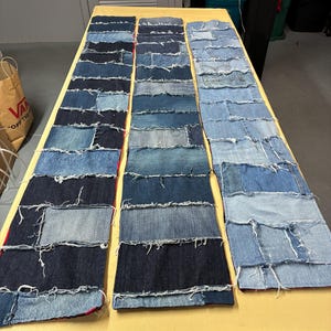 May include: Three denim patchwork runners in varying shades of blue, arranged side-by-side on a yellow surface. Each runner is constructed from rectangular denim pieces, creating a textured, handcrafted appearance. The edges of the denim pieces are frayed.