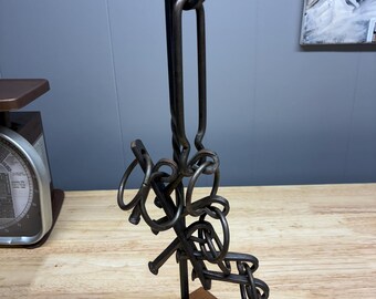 Può includere: Una scultura puzzle in metallo scuro con anelli ad incastro e un elemento verticale, montata su una base quadrata in legno. Il design è intricato e sembra forgiato a mano.