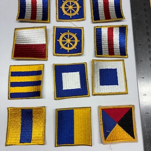 May include: A collection of twelve square embroidered nautical signal flag patches arranged in a grid. Each patch features distinct geometric patterns in blue, white, red, yellow, and black. A metal ruler is visible along the right and bottom edges.