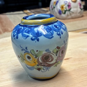 May include: A small, hand-painted ceramic vase with a light blue base. The vase features a yellow rim, blue floral patterns around the top, and colorful roses in yellow, pink, and brown. A second decorative item is visible in the background.