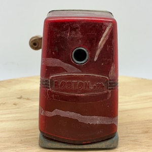 May include: A vintage red Boston pencil sharpener with a wooden crank. The sharpener has the word "BOSTON" embossed on the front. The sharpener sits on a gray base and is set against a light background.