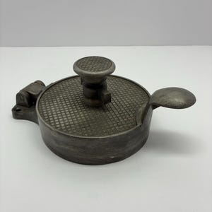 May include: A vintage, circular metal object with a textured surface and a small handle. The object has a round, textured knob on top and a lever on the side. The color is a muted gray.
