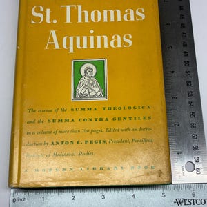 May include: A hardcover book titled Introduction to St. Thomas Aquinas with a yellow cover, a green illustration of a figure, and white text. A metal ruler shows the book is approximately 8.25 inches tall and 5.5 inches wide.