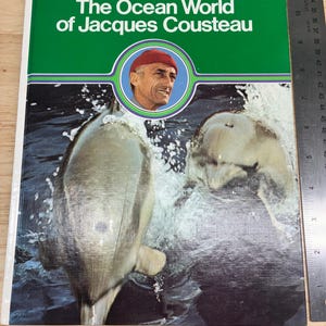 May include: A hardcover book titled The Ocean World of Jacques Cousteau with a green cover featuring a circular portrait of Jacques Cousteau and two dolphins swimming in water. A metal ruler shows the book is approximately 9.5 inches wide.