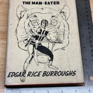 May include: A hardcover book titled Beyond Thirty and The Man-Eater by Edgar Rice Burroughs, featuring a black and white illustration of a figure in front of a large cat face. A metal ruler shows the book is approximately 8.5 inches tall.