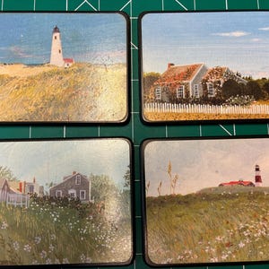 May include: Set of four square coasters with coastal landscape scenes. Each coaster features a different image: a lighthouse, a house with a white picket fence, a house with a flag, and a lighthouse on a hill. The coasters have black edges.