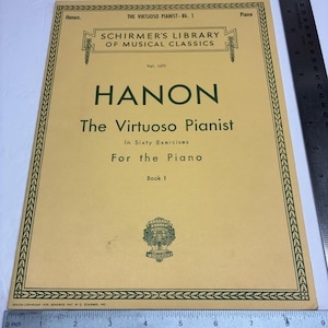 May include: A yellow music book titled Hanon The Virtuoso Pianist in Sixty Exercises for the Piano Book 1, Schirmer's Library of Musical Classics Vol. 1071. A metal ruler shows the book is approximately 9 inches wide and 12 inches tall.