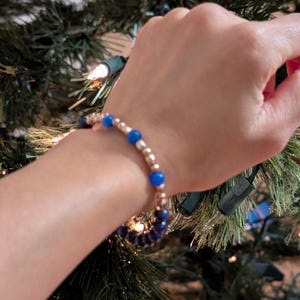May include: A bracelet with blue and gold beads. The bracelet is on a person's wrist, and the background is a Christmas tree with lights.