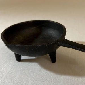May include: A small, black cast iron pan with three legs and a handle. The pan is on a white surface.