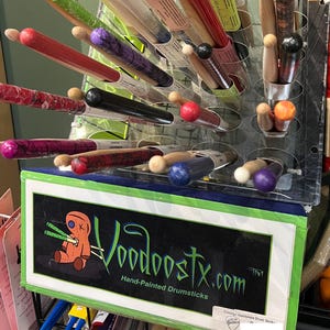 May include: A display rack filled with various hand-painted drumsticks featuring diverse colors and patterns. A sign at the front displays the text Voodoostx.com Hand-Painted Drumsticks with a graphic of an orange voodoo doll holding drumsticks.