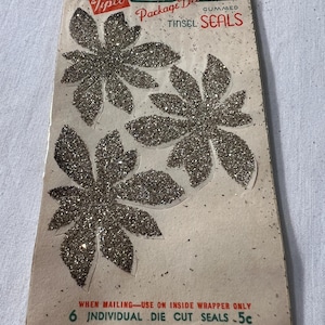 Op de afbeelding: Een verpakking met zes zilveren glitter-uitgesneden zegels in de vorm van bladeren. De verpakking is gelabeld met "Tipco Package Decorations Gummed Tinsel Seals" en "When mailing - use on inside wrapper only 6 individual die cut seals - 5c".