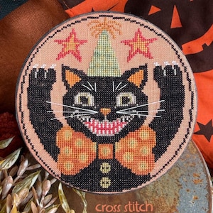 May include: A circular cross stitch pattern featuring a black cat wearing a light green party hat and an orange polka dot bow tie. The cat has white whiskers, a wide grin, and raised paws. Two pink stars flank the hat on a peach background.