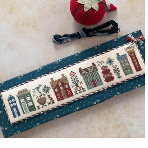 May include: A cross-stitch bookmark featuring a row of colorful houses and floral designs. The bookmark is on a blue patterned surface, with a red strawberry pin cushion and black embroidery floss nearby. The bookmark is approximately 10 inches long.